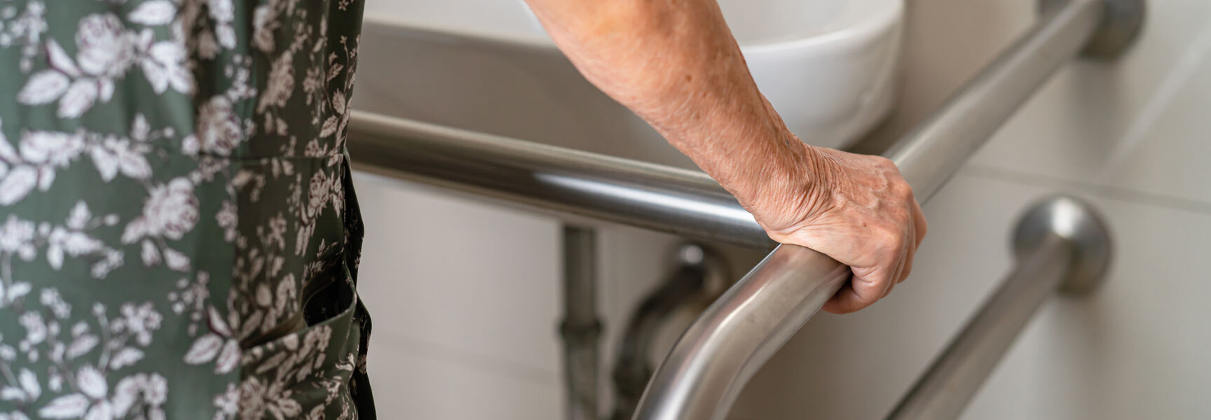 An older woman wearing a floral dress holds onto a metal handle bar for support in a bathroom next to a sink.
