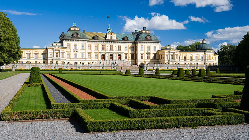 The grand Drottningholm Palace in Sweden, with its manicured gardens and lush green lawns under a bright blue sky with scattered clouds.