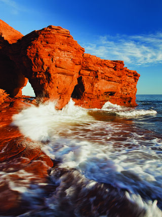 Waves crash against red sandstone cliffs and a natural sea arch along the coastline of Prince Edward Island in the Canadian Maritimes.