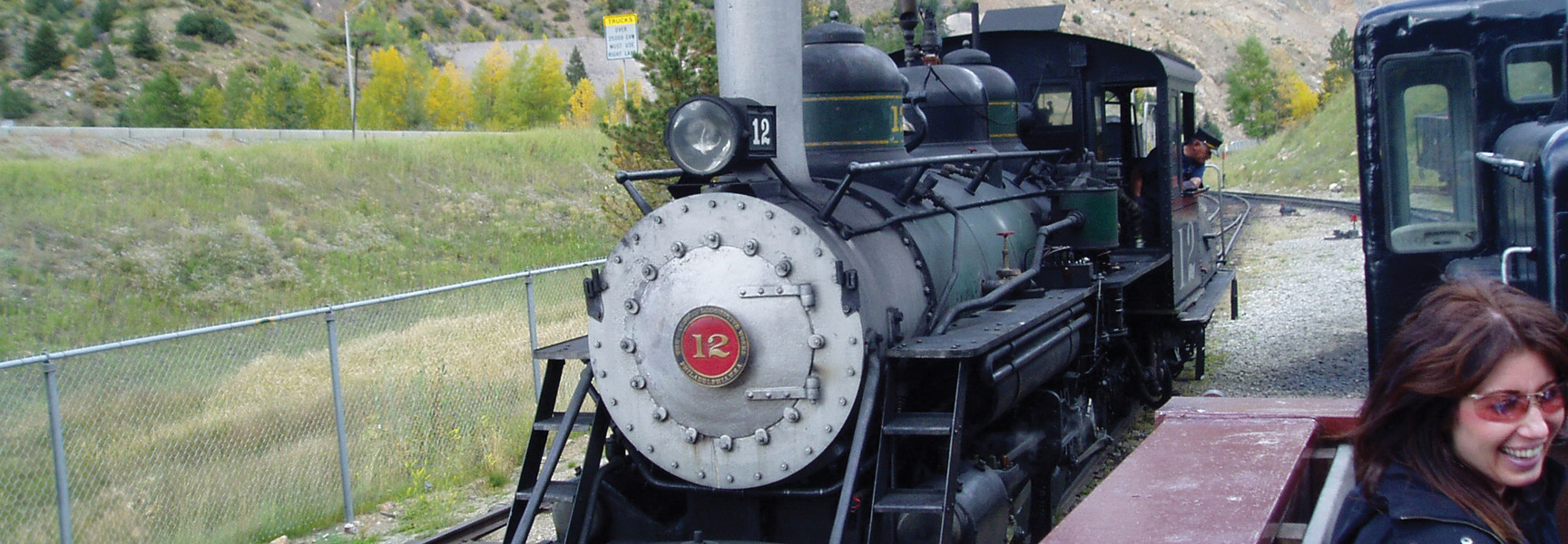 A historic steam engine, number 12, sits on the tracks in the mountains of Colorado, with a smiling woman in the foreground.