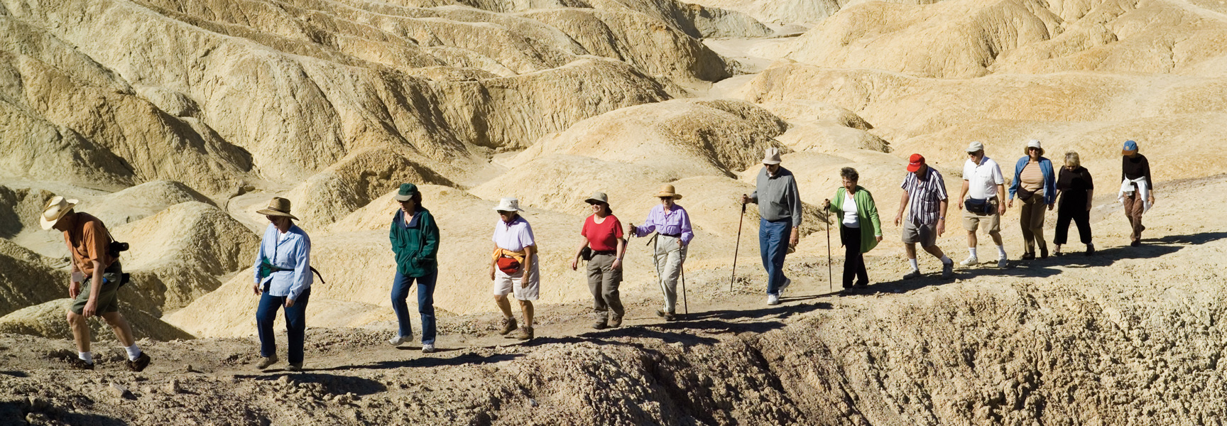 A group of hikers walks in a line on a narrow trail through the golden, rolling badlands of Death Valley National Park, California.