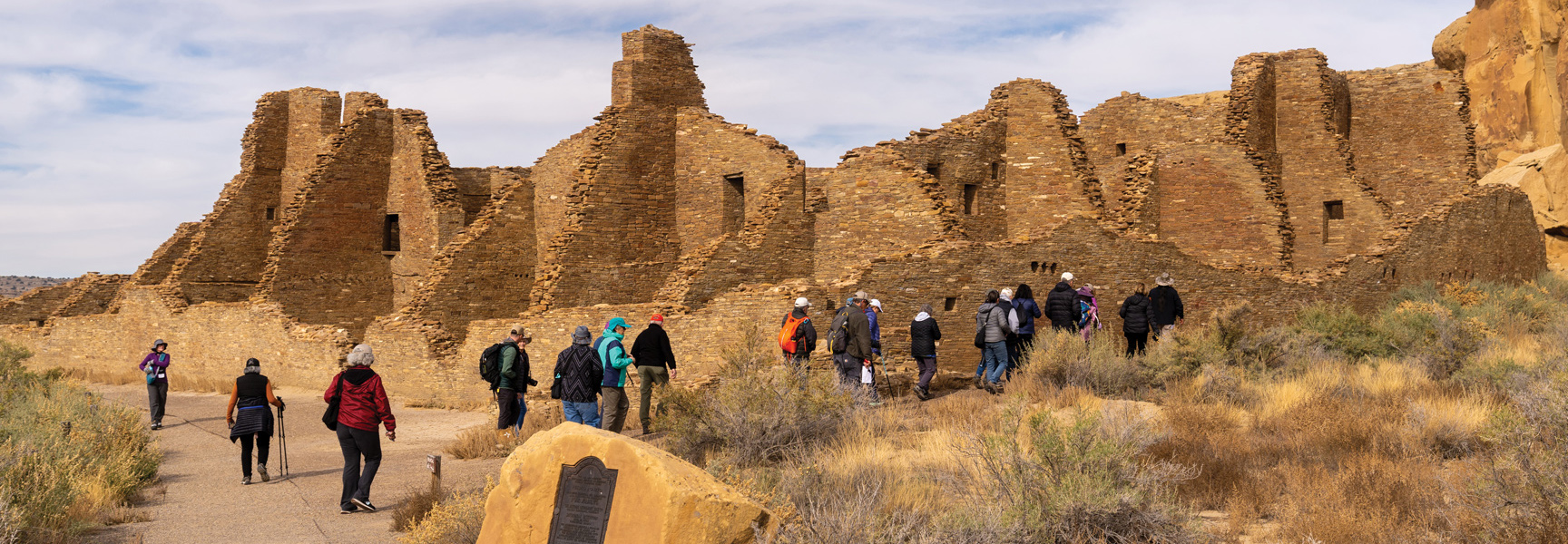 A group of visitors explores the ancient stone ruins of Chaco Canyon in the New Mexico desert on a partly cloudy day.