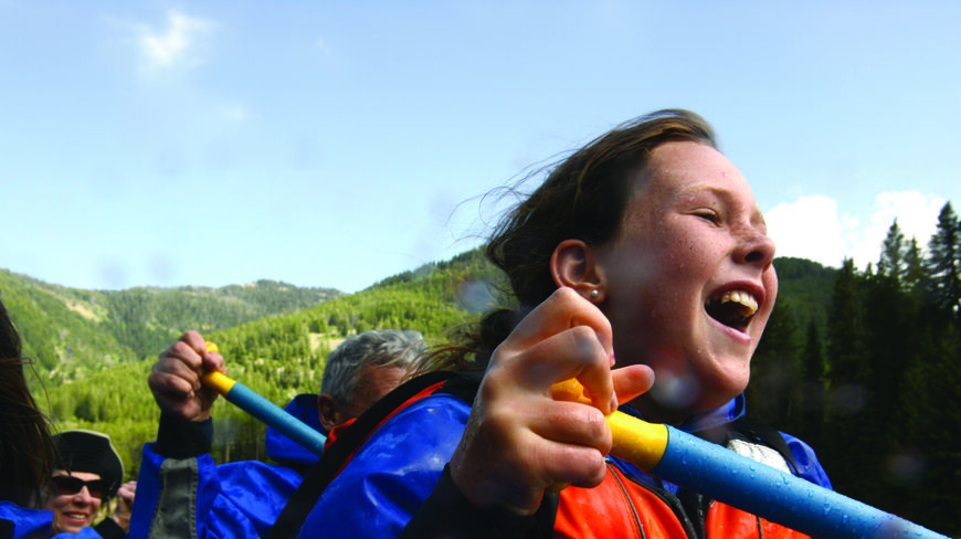 A young grandchild laughs while whitewater rafting on Idaho's Salmon River, holding a blue paddle and wearing an orange life vest amidst forested mountains.