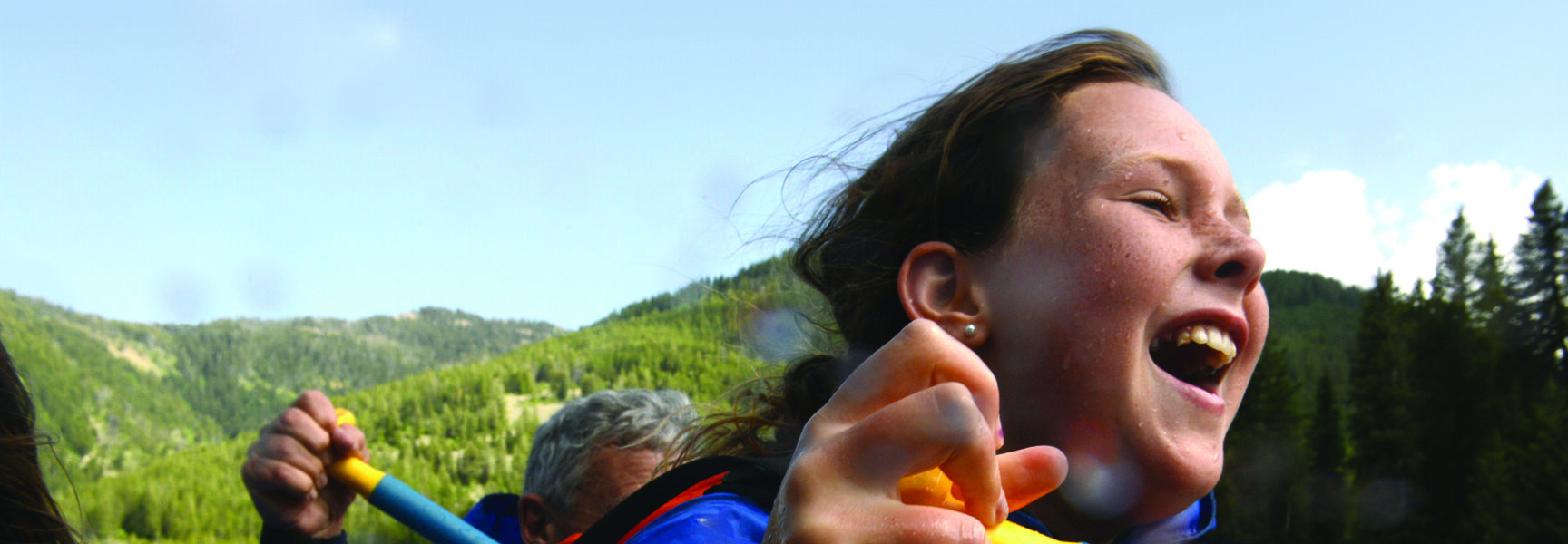 A young grandchild laughs while whitewater rafting on Idaho's Salmon River, holding a blue paddle and wearing an orange life vest amidst forested mountains.