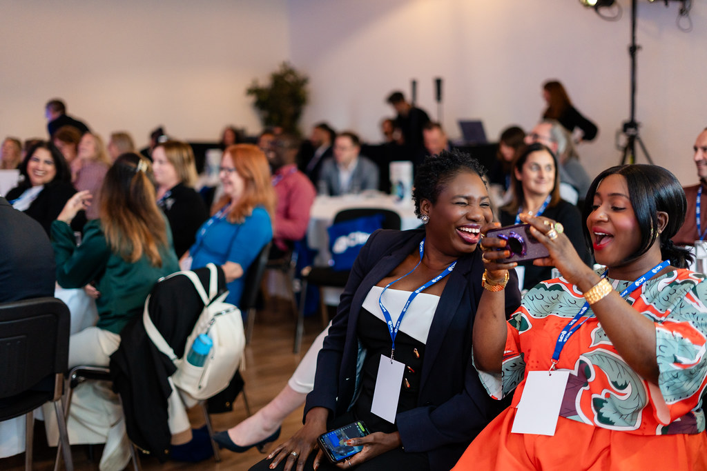 Large group of people sitting at tables looking at stage, two young black women are talking and taking pictures