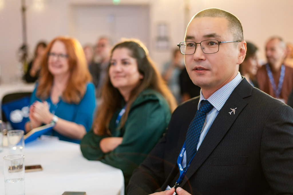 People at conference seated at tables, young man and two women in focus