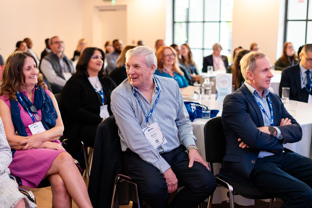 People at conference seated at tables, two middle aged men and one middle aged woman in focus