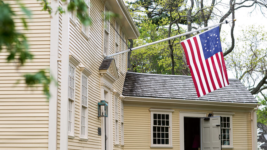 A historic yellow clapboard building in Massachusetts flies a thirteen-star flag, representing a landmark in the birthplace of American liberty.