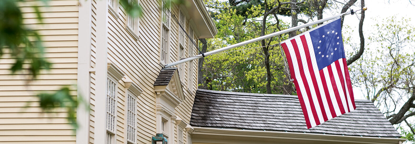 A historic yellow clapboard building in Massachusetts flies a thirteen-star flag, representing a landmark in the birthplace of American liberty.