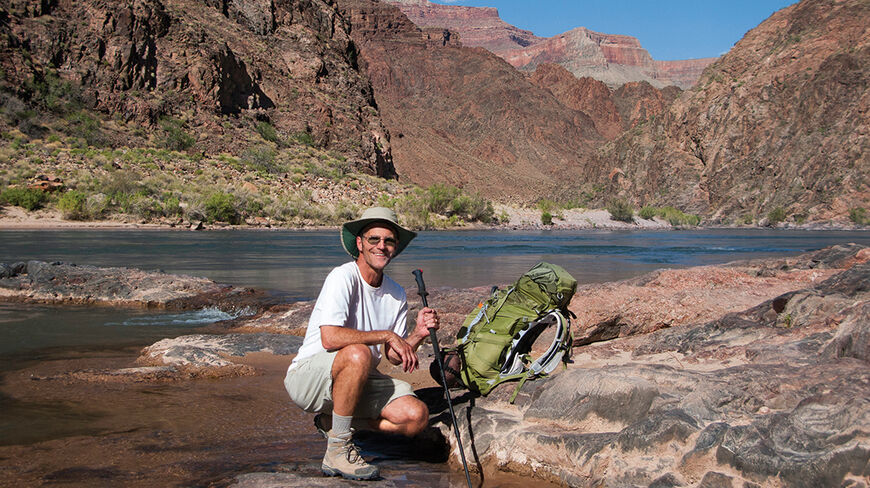 A hiker with a backpack and pole kneels by the river while adventuring below the South Rim of the Grand Canyon in Arizona.