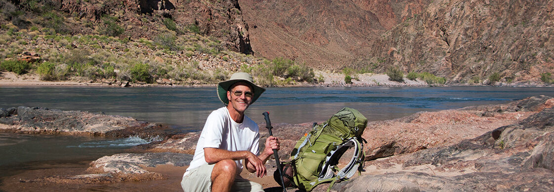 A hiker with a backpack and pole kneels by the river while adventuring below the South Rim of the Grand Canyon in Arizona.