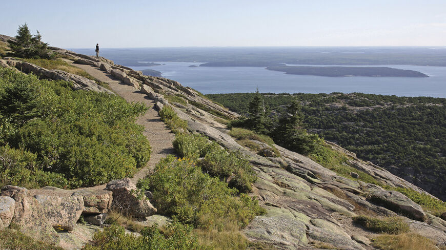 A hiker stands on a rocky trail in Acadia National Park, Maine, overlooking the scenic islands and coastline of Mount Desert Island.