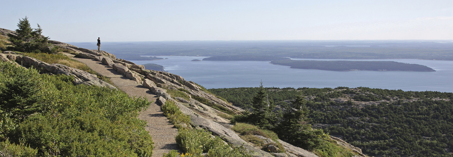 A hiker stands on a rocky trail overlooking the ocean and islands in Acadia National Park, Maine.