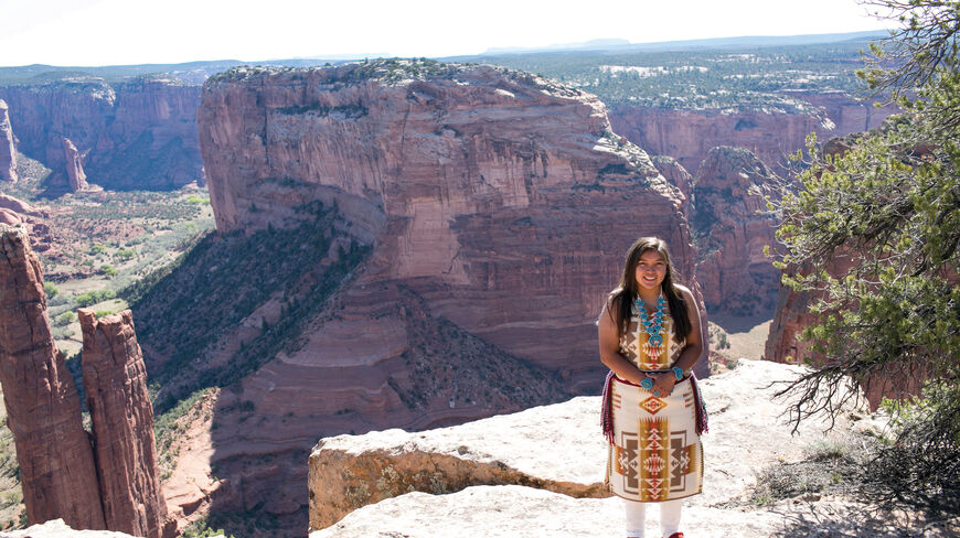 A young Navajo woman in traditional clothing stands on a cliff overlooking a red rock canyon landscape in Arizona.