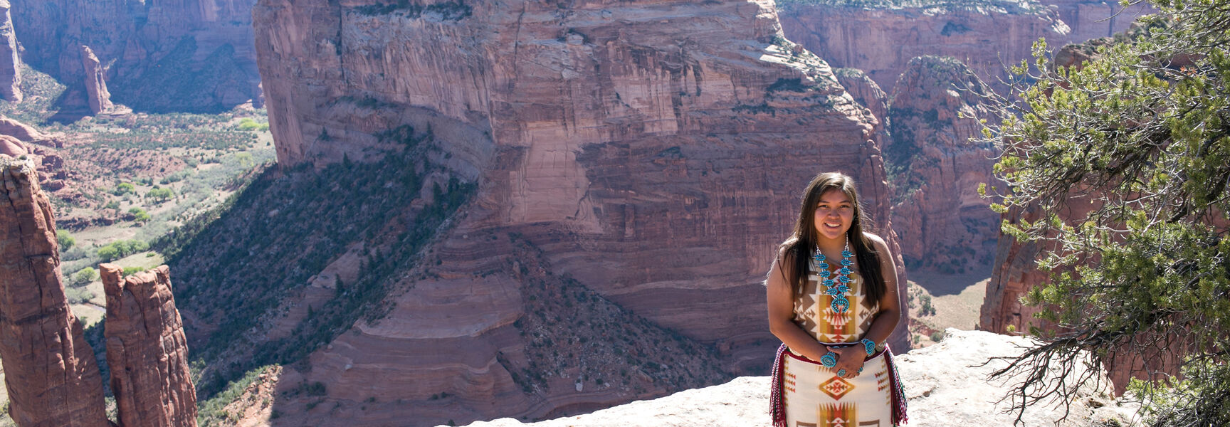 A young Navajo woman in traditional clothing stands on a cliff overlooking a red rock canyon landscape in Arizona.