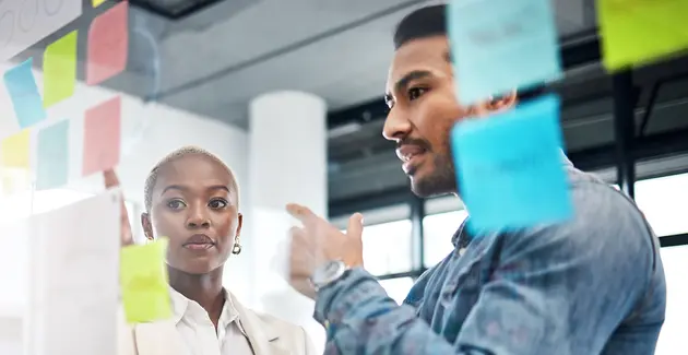 Two professionals, a man and a woman, are engaged in a discussion in an office setting, looking at a glass board covered with colorful sticky notes. The man, on the right and in a casual button-down shirt, is gesturing towards the board as he speaks. The woman, on the left and in professional attire, is looking intently at the board or the man. The sticky notes suggest a brainstorming or project management session.