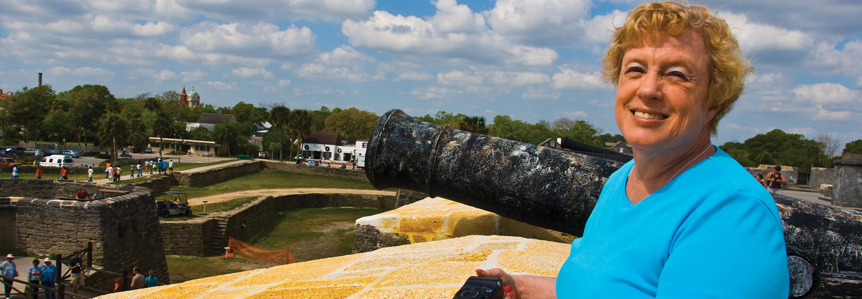 A smiling woman stands next to an old cannon on the walls of a fort, overlooking the city of St. Augustine, Florida.