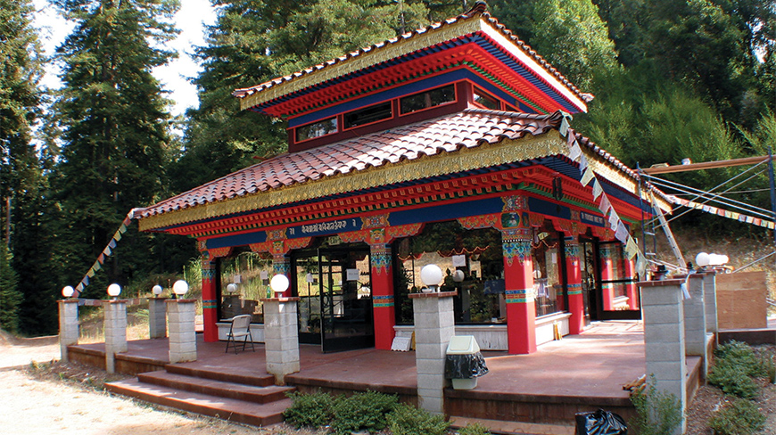 A colorful, traditional Buddhist temple with a tiled roof and prayer flags stands in a California forest.