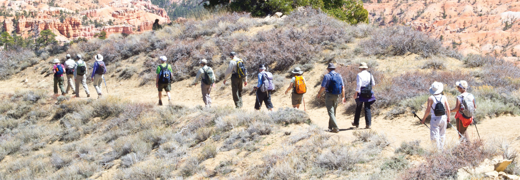 A group of hikers with backpacks walks along a hillside trail in Utah, with red rock formations visible in the background.
