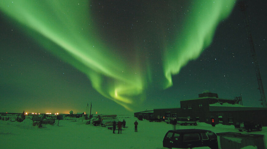 Brilliant green northern lights streak across the starry night sky over a building in the snowy landscape of Churchill, Manitoba.