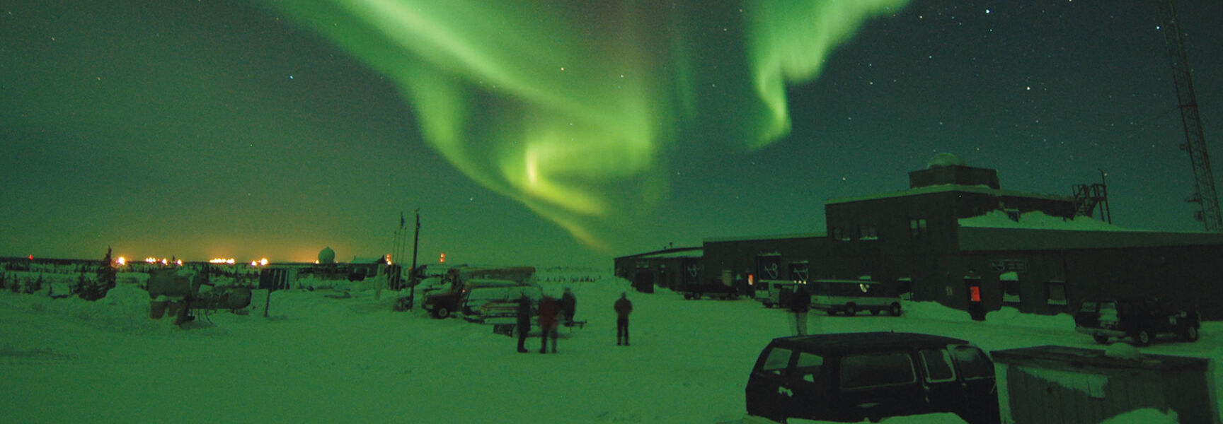 Brilliant green northern lights streak across the starry night sky over a building in the snowy landscape of Churchill, Manitoba.