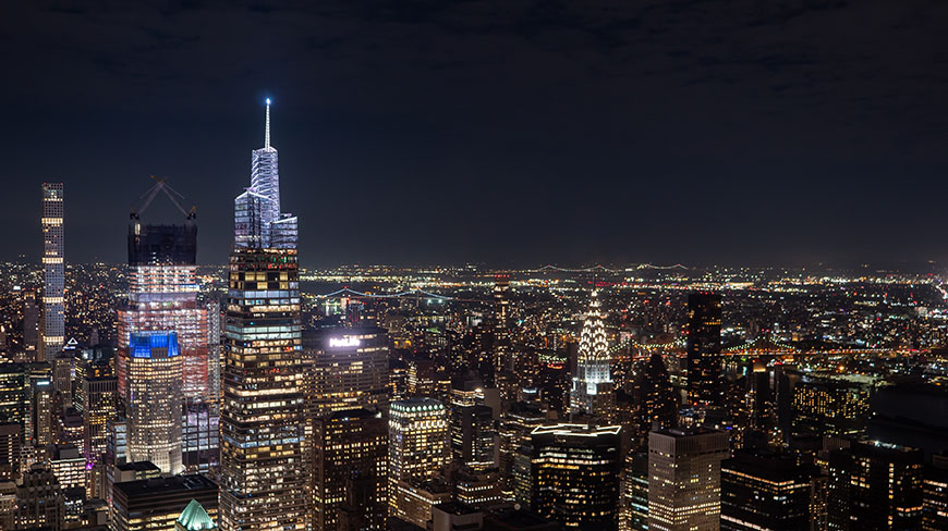 An aerial view of the brightly lit New York City skyline at night, showcasing numerous skyscrapers under a dark sky.