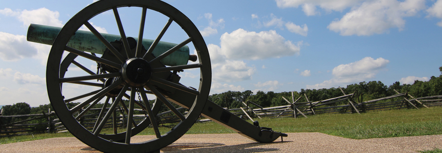 A Civil War cannon rests on the battlefield in Gettysburg, Pennsylvania, with a split-rail fence and trees in the background under a blue sky.