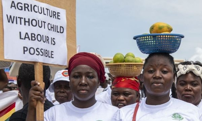 Number of women with fruit baskets balanced on their heads, one of them is holding a sign that reads 'Agriculture without child labour is possible' 