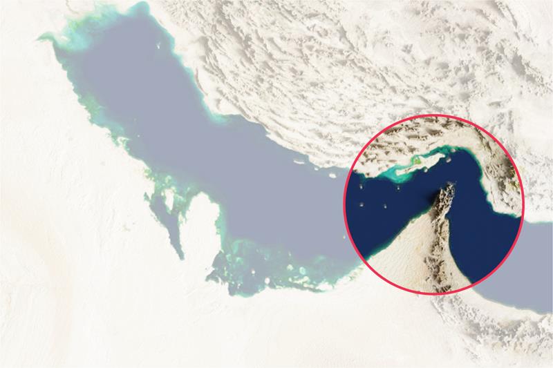 A high-resolution satellite image focusing on the Persian Gulf and the surrounding arid landscape of the Middle East. A red circular inset provides a magnified view of the Strait of Hormuz, highlighting the jagged coastline of the Musandam Peninsula and the narrow waterway between Oman/UAE and Iran.