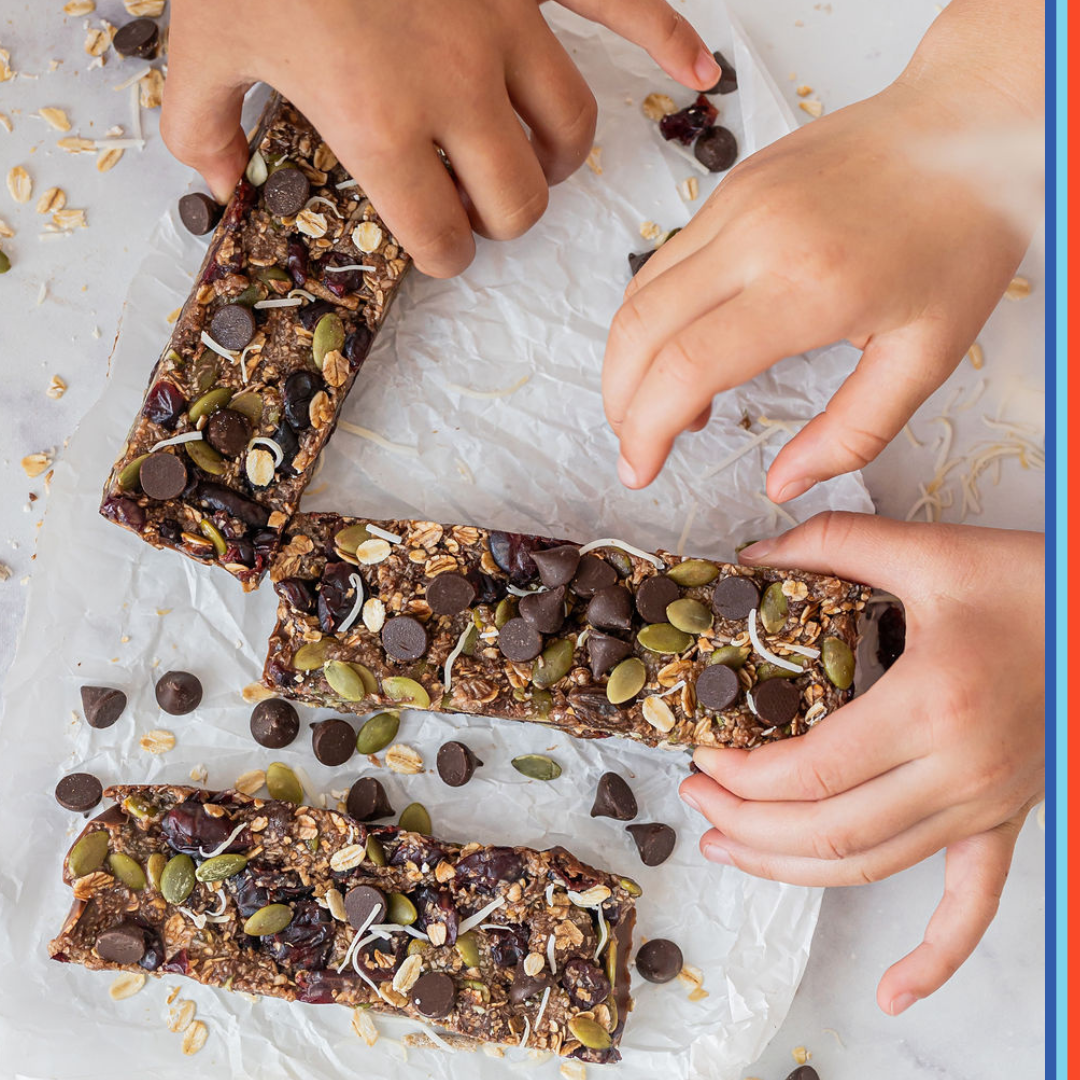 Three homemade granola bars are placed upon baking paper. The bars are sprinkled with pepitas, dark chocolate drops and shredded coconut.