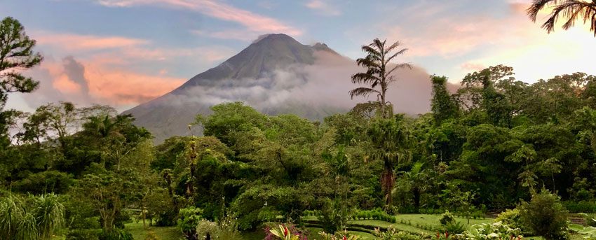 The Arenal Volcano, partially covered in clouds, towers over a lush rainforest and landscaped gardens during a colorful sunset.