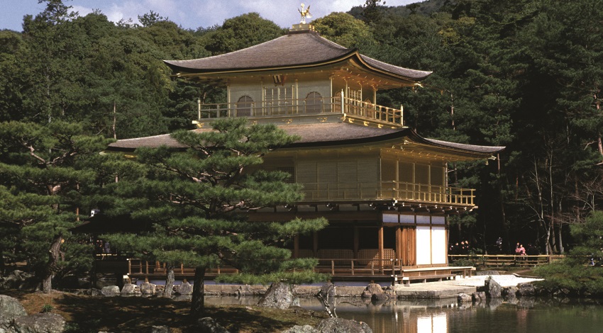 A Zen temple stands by the water with trees in the background and with the sun reflecting off its gold leaf