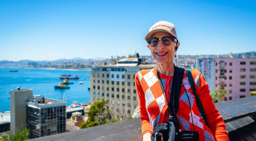 A smiling woman wearing a Road Scholar hat and orange sweater stands before a scenic coastal harbor, illustrating introvert travel tips.