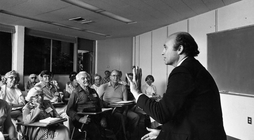 Jim Verschueren speaks and gestures to a classroom of adult students in a black and white photograph.