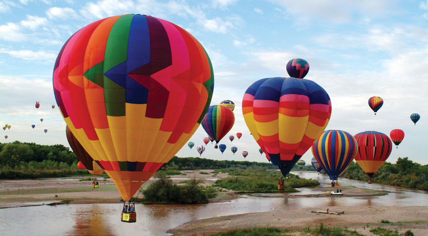 Multiple vibrant hot air balloons drift over a river in Albuquerque, New Mexico, beneath a blue sky filled with light clouds.