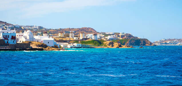 A scenic view of white buildings and windmills on the coastline of Mykonos, Greece, overlooking the deep blue Mediterranean Sea.