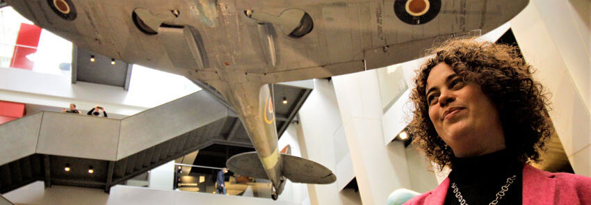 Ruth Polling stands in front of a suspended Spitfire aircraft inside the Imperial War Museum.