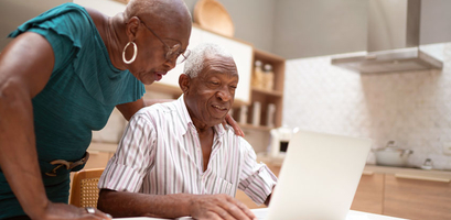 An elderly couple looks at a laptop screen together in their kitchen while researching spring savings.