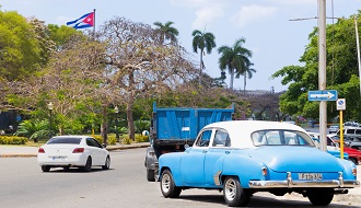 A classic blue car drives along a sunny street in Cuba with a Cuban flag flying in the background.