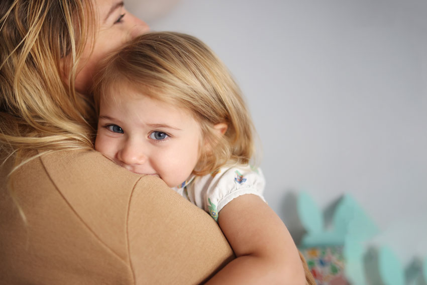 Deklica, ki ima podkožno bulico, se stiska k mami in ji kuka čez ramo.