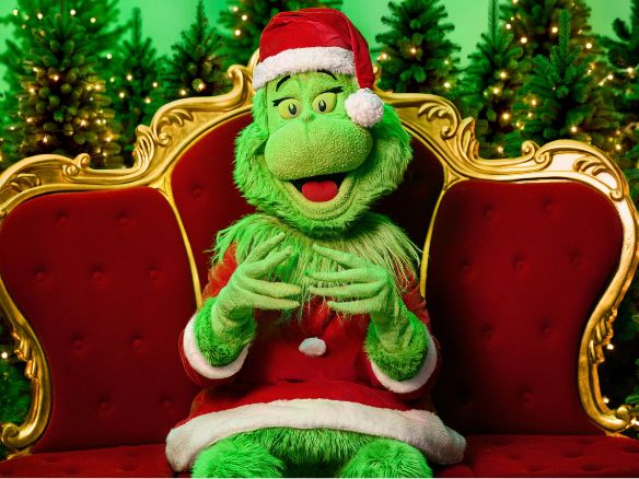 The Grinch in a Santa suit sits cheekily on a red velvet and gold throne, surrounded by lit trees.