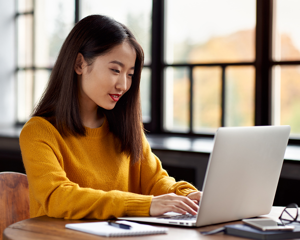 A young woman with dark hair, wearing a mustard yellow sweater, is seated at a wooden table, smiling slightly as she types on a silver laptop. There is a notebook, pen, phone, and glasses nearby, and a large window with an out-of-focus view of trees behind her.