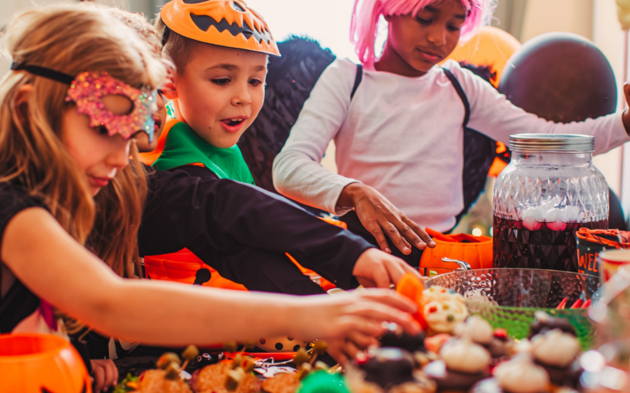 Three children in Halloween costumes gather around a table filled with colorful treats, reaching for food and candy.
