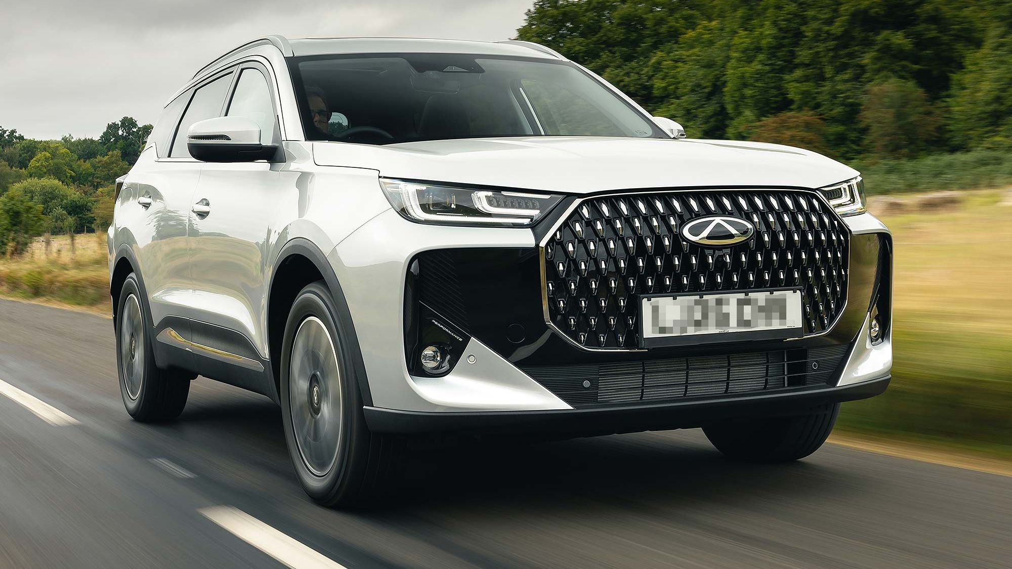 A white Chery Tiggo 7 SUV driving along a country road, front view in motion, available on the Motability Scheme.