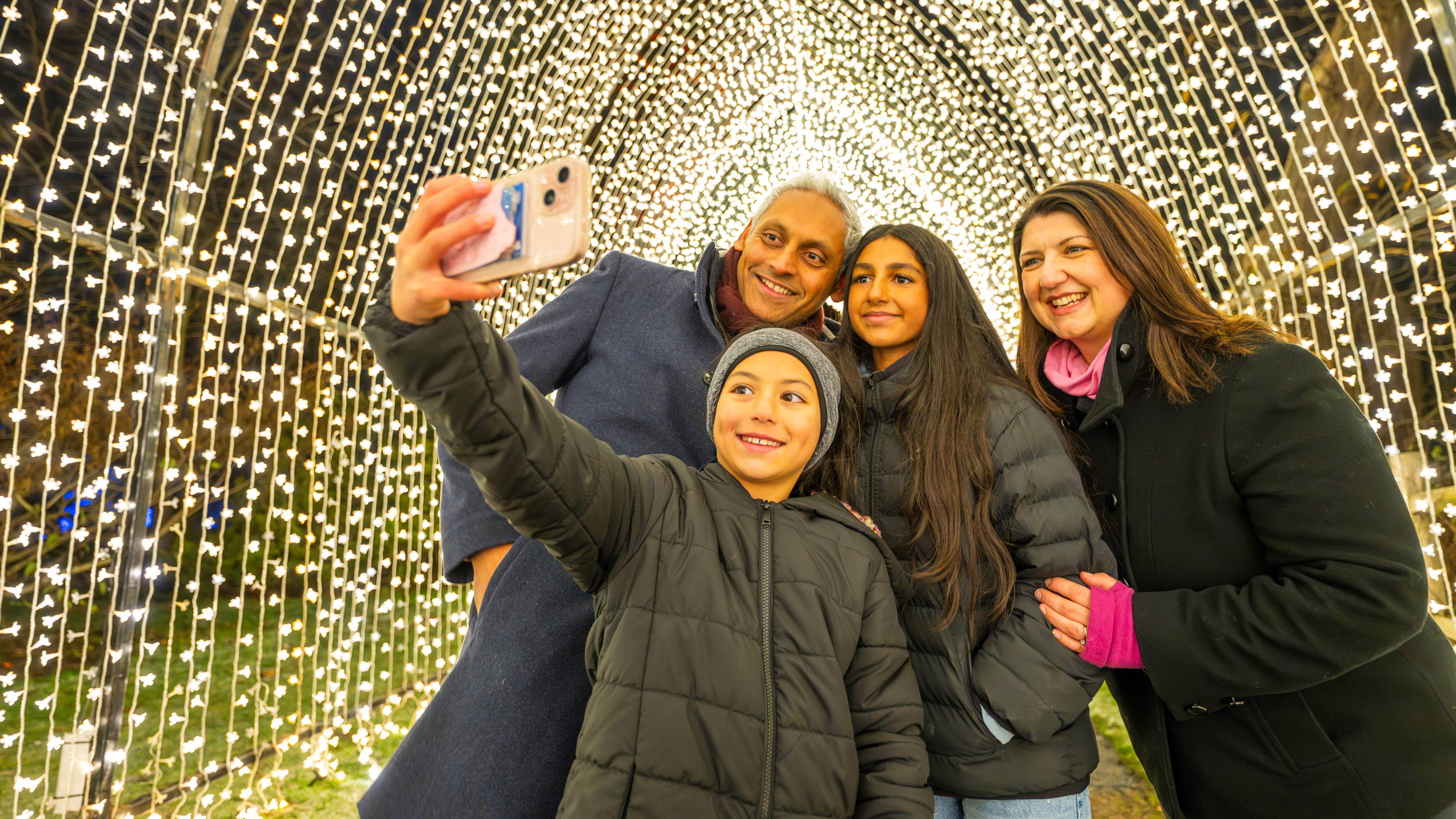 A smiling family of four take a selfie together inside a glowing tunnel of twinkling golden lights at night.