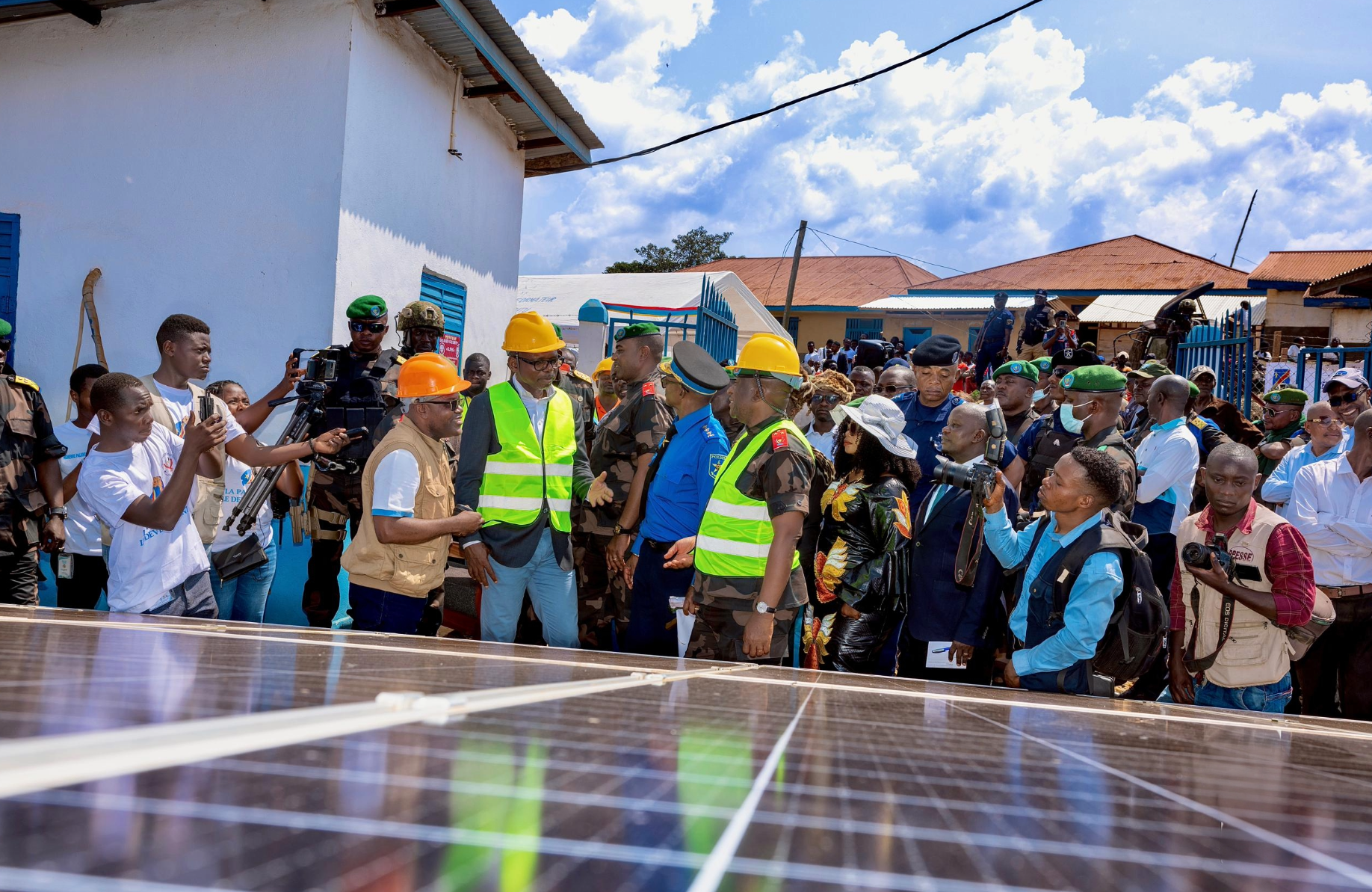 Officials in high-visibility vests and hard hats inspect a solar panel array in the Democratic Republic of Congo, surrounded by a large crowd of security, press, and locals.
