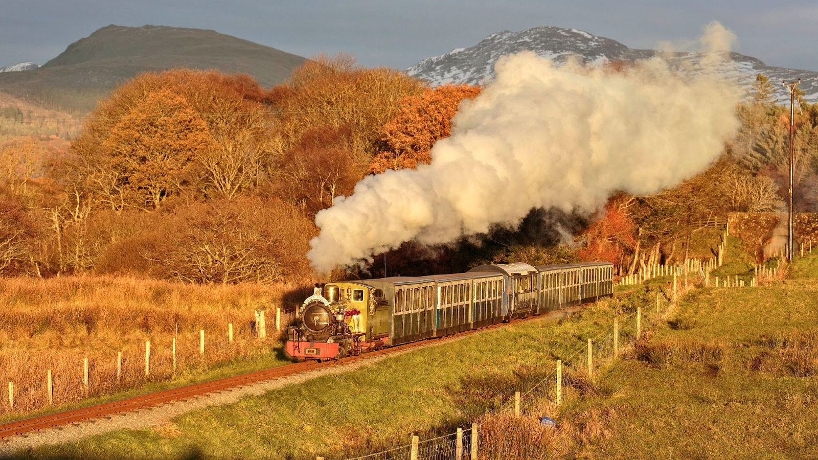 A vintage steam train travels through golden autumn countryside, with smoke billowing from its chimney and snow-dusted hills in the background.