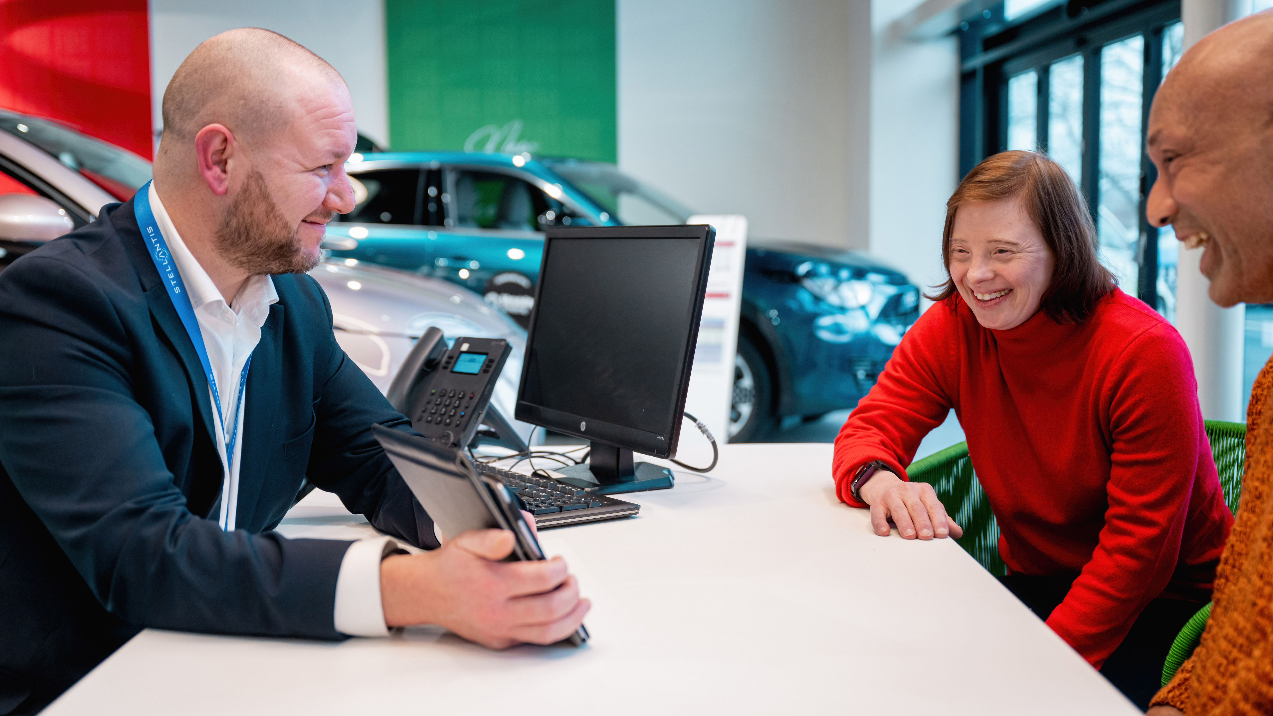 Car dealership staff member sitting at a desk with a tablet, smiling while speaking with two customers across the table, with cars visible in the showroom behind them.