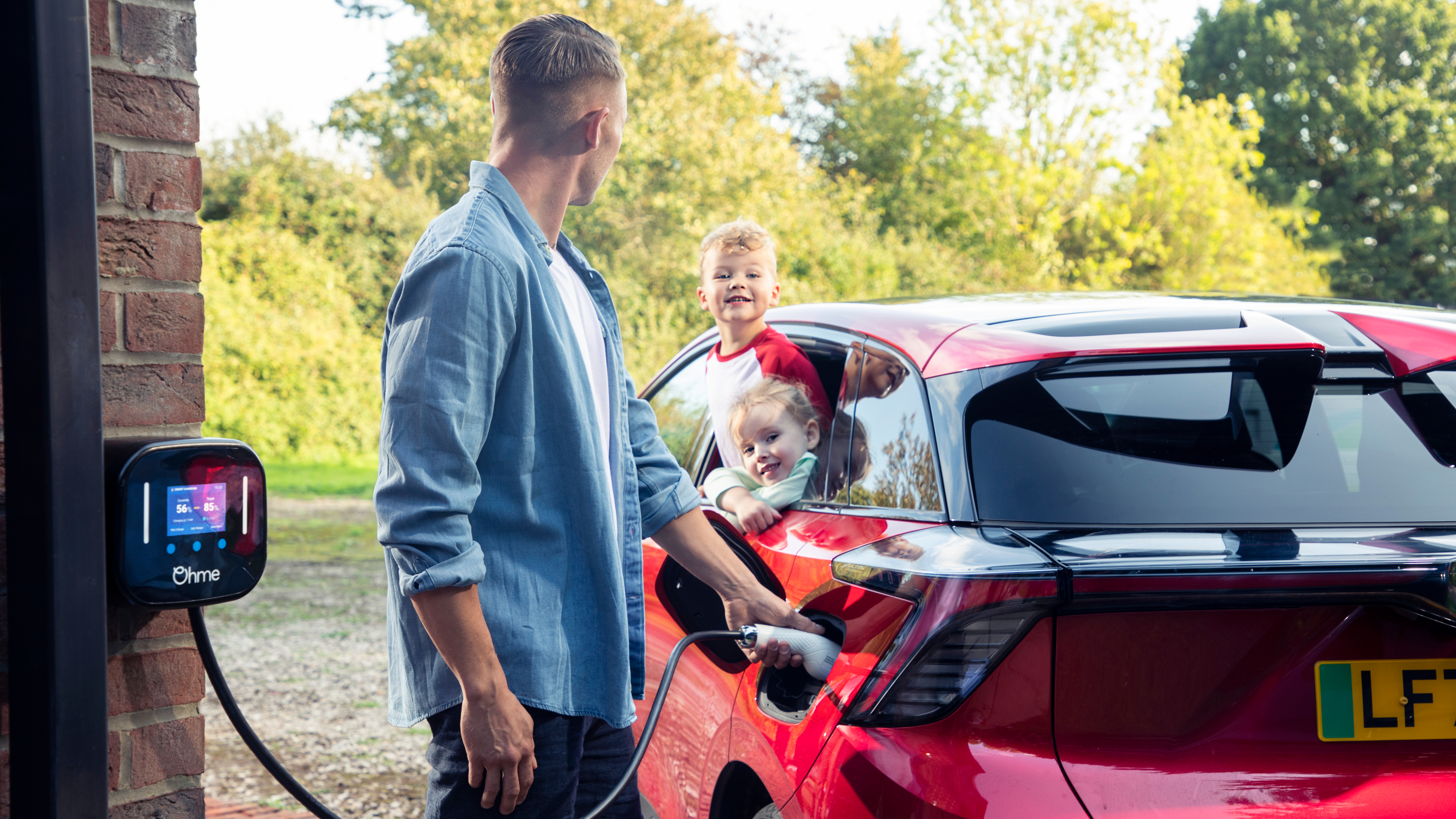 A man charges a red electric car at home using an Ohme wall charger, while two young children smile and lean out of the car window. Trees and greenery are visible in the background.