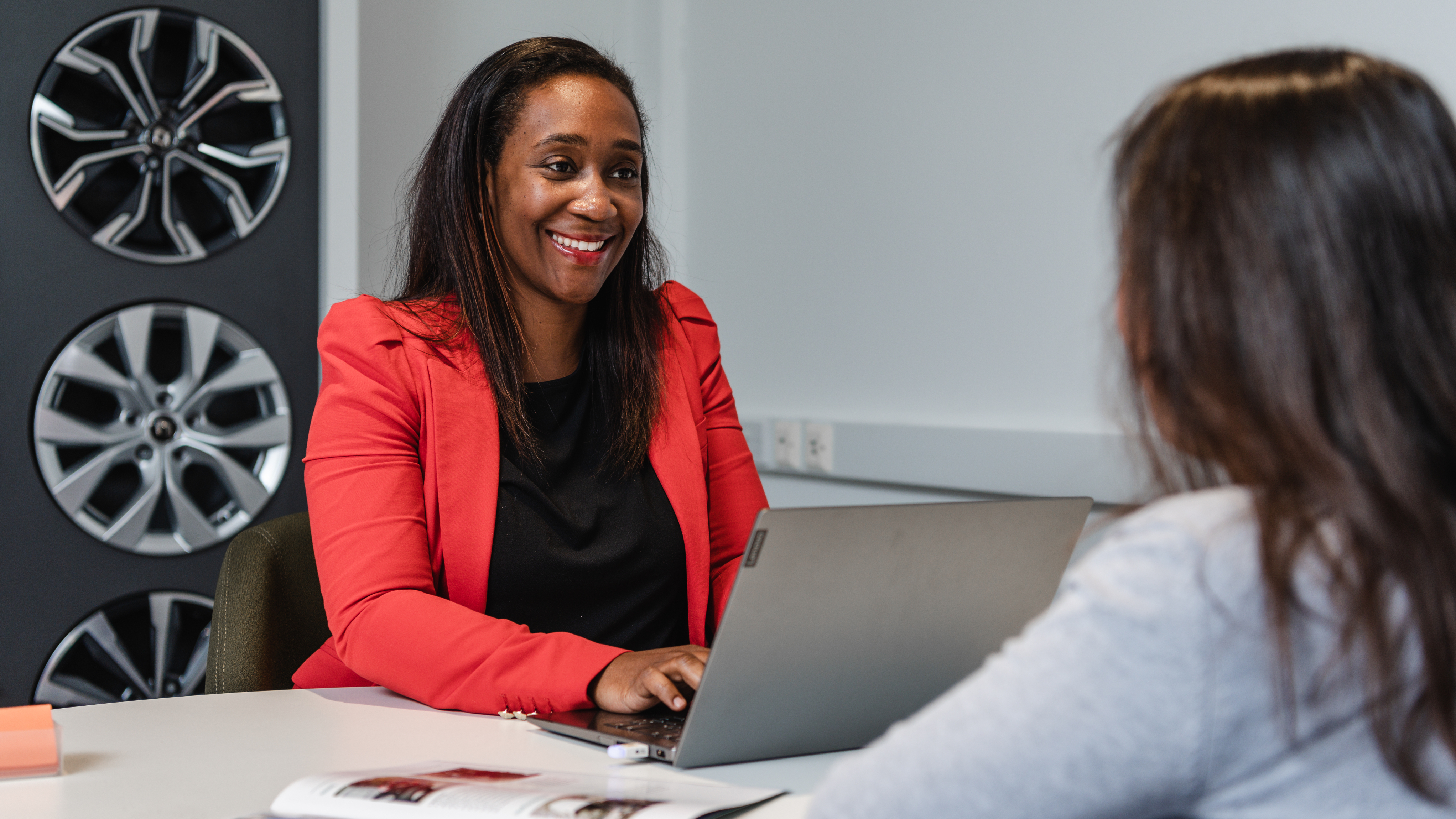 Smiling Motability Scheme car dealer advisor in a red blazer sits at a desk using a laptop while speaking with a customer. Alloy wheel displays are mounted on the wall behind her, and a brochure lies open on the desk between them.